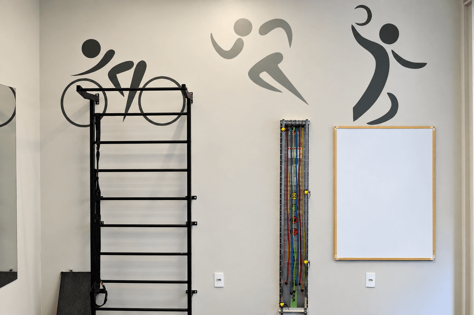 Espaldar de ferro, painel de elásticos e quadro branco em parede com pictogramas de ciclismo, corrida e basquete, na sala funcional da Focus Saúde.
